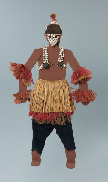 kompletna maska młodej dziewczyny - Ujęcie z przodu. Maska kobiety wykonana z włókien roślinnych. Składa się z „kominiarki” ozdobionej muszlami kauri nakładanej na głowę, napierśnika z miseczkami wykonanymi z owoców baobabu, również ozdobionego muszlami kauri i różnobarwnymi szklanymi paciorkami, bawełnianych spodni samodziałowych barwionych indygo, trzech spódniczek oraz bransolet nakładanych na ramiona i nadgarstki.