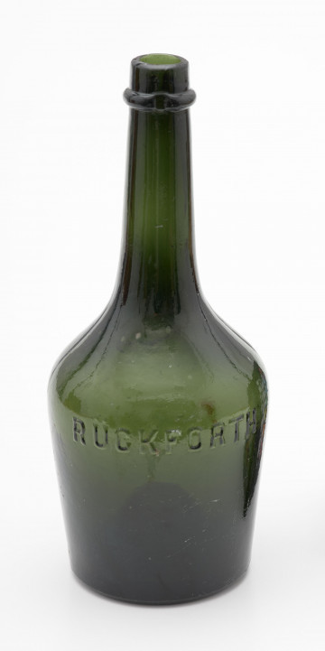 szklana butelka z korpusem rozszerzającym się ku górze - Ujęcie z przodu; Szklana butelka piwówka z grubego szkła butelkowego w ciemnym kolorze oliwkowo-zielonym. Korpus niski, gruszkowy, ponad stopą lekko rozszerzający się ku górze, a następnie silnie zwężający i przechodzący w szyjkę. Poniżej wylewu szyjki pogrubiony pierścień mieszczący dwa zagłębienia na umieszczenie zamknięcia. Dno silnie wklęsłe.