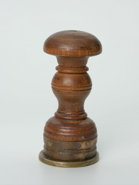 tłok pieczętny - Ujęcie z boku; Okrągły tłok pieczętny z brązu z drewniano-rogowym uchwytem oraz rysunkiem produktów rzemieślniczych na tarczy w polu pieczęci.