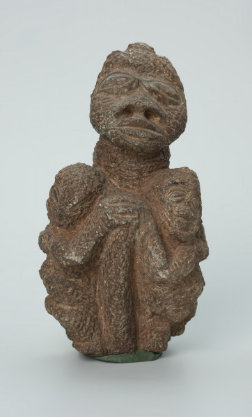 rzeźba; przedmiot obrzędowy; figura kultu zmarłych - Ujęcie z przodu. Rzeźbiona w ziemistym steatycie siedząca postać ludzka o cechach kobiecych, otoczona przez trójkę mniejszych, dzieciecych postaci, skierowanych ku niej z uniesionymi rękoma.