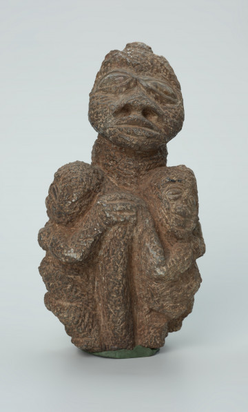 rzeźba; przedmiot obrzędowy; figura kultu zmarłych - Ujęcie z przodu. Rzeźbiona w ziemistym steatycie siedząca postać ludzka o cechach kobiecych, otoczona przez trójkę mniejszych, dzieciecych postaci, skierowanych ku niej z uniesionymi rękoma.