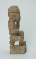 figura kultu przodków - Ujęcie z boku z prawej. Ustawiona na kwadratowej podstawie, wyrzeźbiona w biało-szarym kamieniu postać zamyślonego, siedzącego na stołku mężczyzny w czapce, o zaokrąglonych rogach i spłaszczonej części górnej.