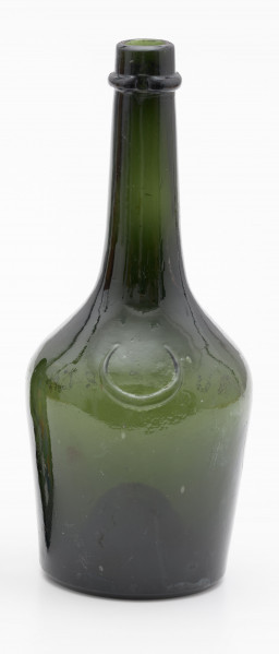 szklana butelka z korpusem rozszerzającym się ku górze - Ujęcie z tyłu; Butelka piwówka z grubego szkła butelkowego w ciemnym kolorze oliwkowo-zielonym. Korpus niski, gruszkowy, ponad stopą lekko rozszerzający się ku górze, a następnie silnie zwężający i przechodzący w szyjkę. Poniżej wylewu szyjki pogrubiony pierścień mieszczący dwa zagłębienia na umieszczenie zamknięcia. Dno silnie wklęsłe.