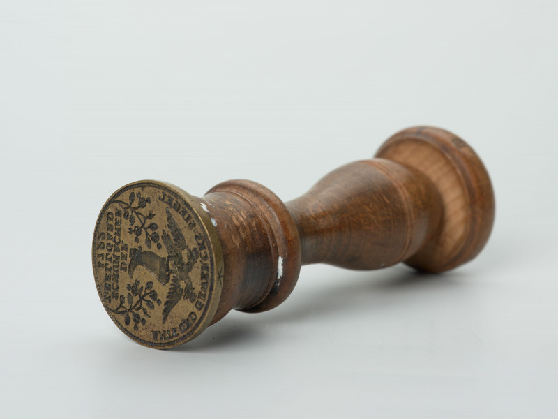 tłok pieczętny - Ujęcie z dołu; Okrągły tłok pieczętny z brązu z drewnianym uchwytem oraz rysunkiem buta z dwugłowym orłem w polu pieczęci.