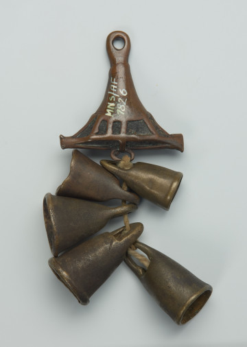 biżuteria, ozdoba - Ujęcie z przodu. Mały, trójkątny, ozdobiony wzorami geometrycznymi wisiorek, wykonany z brązu, z którego zwisa pięć dzwoneczków umieszczonych na bawełnianym sznurku.