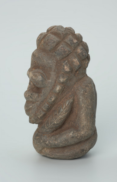 rzeźba; przedmiot obrzędowy; Figura kultu zmarłych - Ujęcie lewy bok. Rzeźbiona w szarobeżowym steatycie siedząca postać ludzka o cechach męskich, w prawej dłoni trzymajaca przedmiot przypominający grot, a w lewej pióro lub liść.