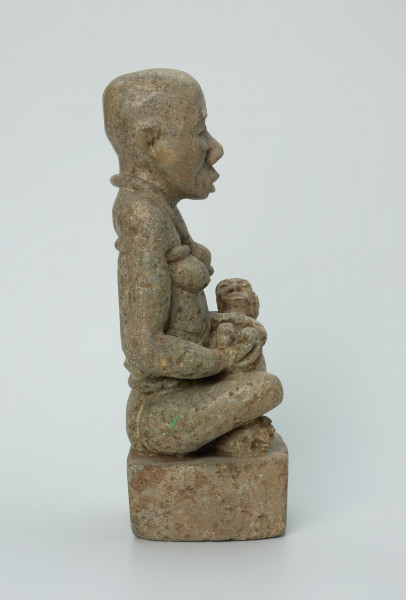 figura kultu przodków - Ujęcie z boku z prawej. Rzeźbiona w biało-szarym kamieniu dość duża postać kobiety w pozycji siedzącej z nogami skrzyżowanymi. Kobieta siedzi na małej, kwadratowej, lekko zaokrąglonej na rogach podstawie, w dłoniach trzyma dziecko. Widoczne rysy, mikropęknięcia i liczne malutkie otworki.