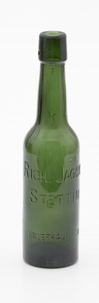 szklana butelka w ciemnym kolorze oliwkowo-zielonym - Ujęcie z przodu; Szklana butelka piwówka z grubego szkła butelkowego w ciemnym kolorze oliwkowo-zielonym. Szyjka wyodrębniona z pogrubieniem przy wylewie, u dołu pogrubienia z dwoma otworami do zamknięcia pałąkowego. Dno zagłębione płaskie. Ślad szwóww bocznych.