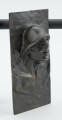 portret męski - Ujęcie z przodu skosem w prawą stronę; Płaskorzeźba przedstawiająca głowę mężczyzny. Z blachy stanowiącej tło wyłania się kwadratowa twarz z szerokim nosem i rozchylonymi ustami, szyja mocno osadzona, szeroka. Na głowie hutniczy kask. Widoczny fragment torsu.