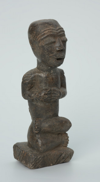 Ujęcie z przodu z prawej. Rzeźbiona w grafitowym kamieniu postać ludzka w pozycji siedzącej - najprawdopodobniej mężczyzna. Rzeźba ma charakterystyczne nacięcia - skaryfikacje wykonane na ramionach oraz plecach. Widoczne rysy i mikropęknięcia.