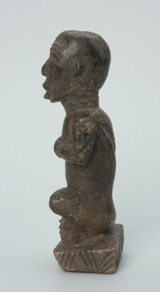 figura kultu przodków - Ujęcie z boku z lewej. Rzeźbiona w grafitowym kamieniu postać ludzka w pozycji siedzącej - najprawdopodobniej mężczyzna. Rzeźba ma charakterystyczne nacięcia - skaryfikacje wykonane na ramionach oraz plecach. Widoczne rysy i mikropęknięcia.