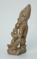 Ujęcie z boku z lewej. Rzeźbiona w biało-szarym kamieniu postać mężczyzny palącego fajkę w pozycji siedzącej. Mężczyzna siedzi na okrągłym, malutkim stołku, ma charakterystyczne nakrycie głowy - spiczastą, ozdobioną łączącymi się ukośnymi nacięciami czapkę o zaokrąglonych rogach i pomponiku. Widoczne rysy, mikropęknięcia, liczne malutkie otworki i przebarwienia.