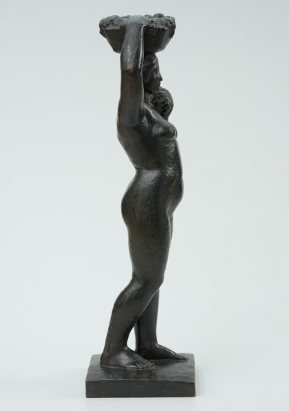 akt kobiecy - Ujęcie z boku z prawej strony; Posążek przedstawia postać stojącej kobiety o pełnych kształtach dźwigającą kosz na głowie. Sylwetka wyprostowana o masywnych nogach, biodrach i niewielkich piersiach, zupełnie naga, z okrągłą twarzą i krótkich włosach- prawa noga lekko cofnięta, lewa ręka zgięta w łokciu dzierży na wysokości barku kiść owoców, prawa uniesiona ręka przytrzymuje na głowie płaski okrągły niski kosz wypełniony owocami. Figurka stoi na kwadratowej niskiej podstawce.