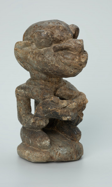 rzeźba; przedmiot obrzędowy; Figura kultu sił wegetacji - Ujęcie z przodu z prawej strony. Rzeźbiona w białoszarym steatycie siedząca na płaskiej, owalnej podstawie kobieta trzymająca w dłoniach, przy biuście zwrócone ku niej dziecko.