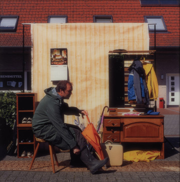 fotografia - Ujęcie z przodu. Fotografia barwna przedstawiająca mężczyznę siedzącego w zaaranżowanym w przestrzeni miejskiej przedpokoju. W tle domki jednorodzinne.