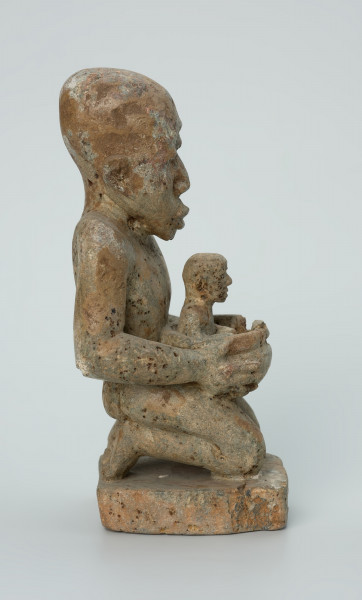 Ujęcie z boku z prawej. Rzeźbiona w biało-szarym kamieniu postać kobiety w pozycji klęczącej. Kobieta trzyma w naczyniu dziecko. Widoczne rysy, mikropęknięcia, liczne malutkie otworki i przebarwienia.