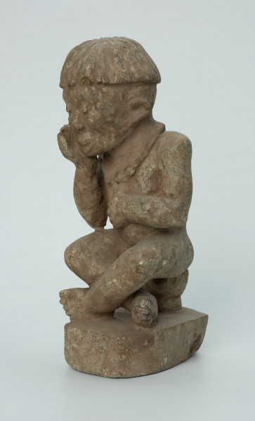 Ujęcie z przodu z lewej. Rzeźbiona w biało-szarym kamieniu postać mężczyzny w pozycji siedzącej. Mężczyzna siedzi na okrągłym, malutkim stołku, ma charakterystyczne nakrycie głowy - czapkę modelowaną promieniście oraz naszyjnik. Widoczne rysy, mikropęknięcia i malutkie otworki.