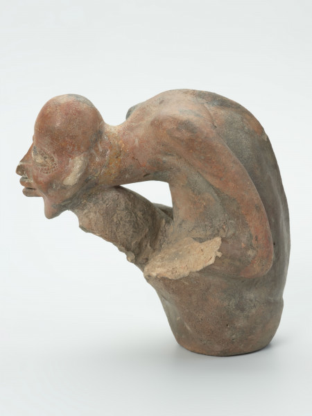 figura - Ujęcie z lewej strony; Gliniana rzeźba postaci ludzkiej w kolorze beżowo-brązowo-ceglastym. Postać w pozycji siedzącej, mocno pochylona do przodu. Twarz oparto na prawej dłoni, która usytuowana została na prawym kolanie. Rzeźbie nie zachowały się w całości nogi (prawa poniżej kolana, lewa poniżej uda) oraz lewe przedramię i dłoń. Postaci zaznaczono bardzo wyraźną linię kręgosłupa.