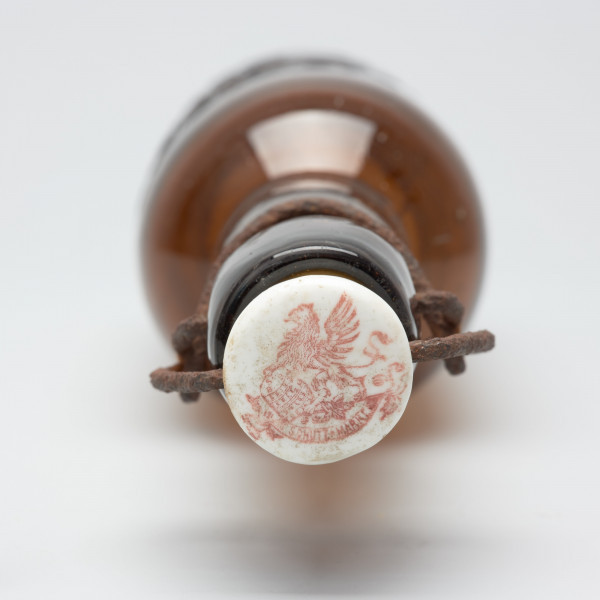 szklana butelka z wyodrębnioną szyjką - Ujęcie z góry; Kapsel z białego porcelitu na pałąku z drutu żelaznego.  Na nim pieczęć czerwona z orłem.