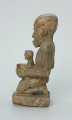 Ujęcie z boku z lewej. Rzeźbiona w biało-szarym kamieniu postać kobiety w pozycji klęczącej. Kobieta trzyma w naczyniu dziecko. Widoczne rysy, mikropęknięcia, liczne malutkie otworki i przebarwienia.