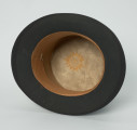 czarny cylinder z welurowej tkaniny - Ujęcie wnętrza; Czarny cylinder z welurowej tkaniny z czarnym otokiem ponad rondem wykończonym pasmanterią. Wewnątrz skórkowy otok. Emaliowana literka 