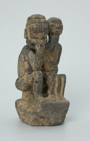 Ujęcie z przodu z prawej. Rzeźbione w grafitowym kamieniu dwie złączone ze sobą postaci ludzkie w pozycji siedzącej. Stykają się ramionami, tułowiem oraz nogami. Jedna z nich – najprawdopodobniej mężczyzna - trzyma w prawej dłoni fajkę. Rzeźba pokryta jest piaskiem. Widoczne rysy, mikropęknięcia, malutkie otworki, przebarwienia oraz ślady klejenia w okolicach szyi.