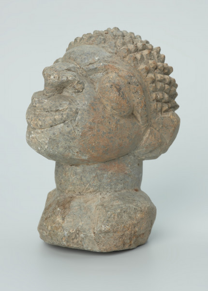 rzeźba; głowa; figura kultu zmarłych - Ujęcie z przodu z lewej strony. Rzeźbiona w szarobeżowym steatycie, usadowiona na okrągłej podstawce, głowa postaci ludzkiej z dużymi uszami z tyłu i w charakterystycznym uczesani w formie małych, lekko zaokrąglonych stożków, być może loczków.
