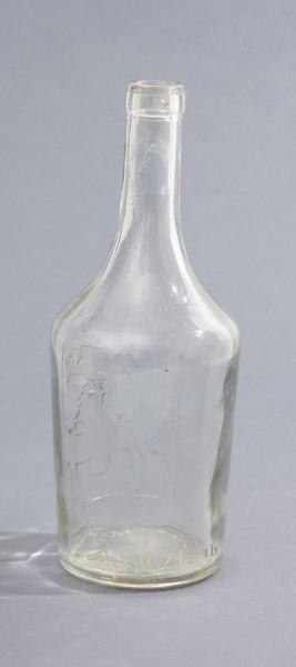 szklana butelka z szerokim korpusem - Ujęcie z tyłu; Butelka piwa z grubego półbiałego przeźroczystego szkła. Korpus szeroki, lekko zwężający się ku dołowi, a od góry przechodzący w szyjkę zakończoną płaskimi pierścieniem. Czytelne szwy boczne.