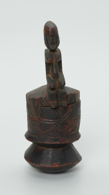 drewninany pojemnik na tytoń z pokrywką - Ujęcie z przodu. Drewniany pojemnik na tytoń z pokrywą, na której znajduje się rzeźbiona, siedząca postać.
