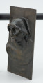 portret męski - Ujęcie z przodu skosem w lewą stronę; Płaskorzeźba przedstawiająca głowę mężczyzny. Z blachy stanowiącej tło wyłania się kwadratowa twarz z szerokim nosem i rozchylonymi ustami, szyja mocno osadzona, szeroka. Na głowie hutniczy kask. Widoczny fragment torsu.