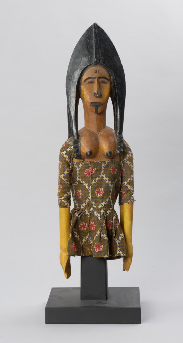 lalka teatralna: Djinè Faro (bóstwo Faro) - Ujęcie z przodu. Drewniana lalka przedstawiająca postać kobiecą o jasnej cerze. Twarz pociągła, nos długi, duże usta, ozdobne nakrycie głowy w kolorze czarnym. Ubrana we wzorzystą sukienkę odsłaniającą nagie piersi.
