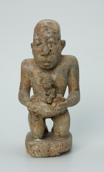 Ujęcie z przodu. Rzeźbiona w biało-szarym kamieniu postać kobiety w pozycji klęczącej. Kobieta trzyma w naczyniu dziecko. Widoczne rysy, mikropęknięcia, liczne malutkie otworki i przebarwienia.