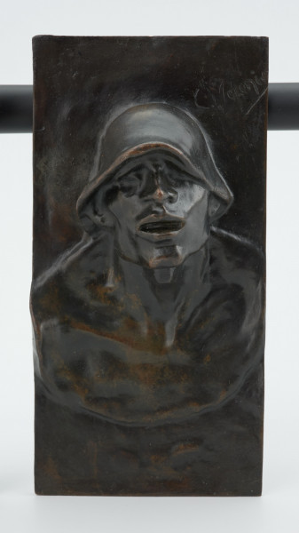 portret męski - Ujęcie z przodu; Płaskorzeźba przedstawiająca głowę mężczyzny. Z blachy stanowiącej tło wyłania się kwadratowa twarz z szerokim nosem i rozchylonymi ustami, szyja mocno osadzona, szeroka. Na głowie hutniczy kask. Widoczny fragment torsu.
