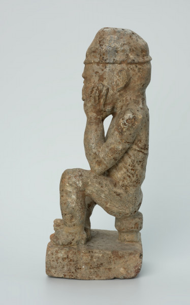 figura kultu przodków - Ujęcie z boku z lewej. Ustawiona na kwadratowej podstawie, wyrzeźbiona w biało-szarym kamieniu postać zamyślonego, siedzącego na stołku mężczyzny w czapce, o zaokrąglonych rogach i spłaszczonej części górnej.