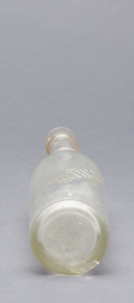szklana butelka z wyodrębnioną szyjką z trzema pierścieniami - Ujęcie z dołu; Szklana butelka piwówka z grubego szkła butelkowego półbiałego przezroczystego. Szyjka wyodrębniona trzema wypukłymi, wąskimi pierścieniami, umieszczonymi w dolnej części szyjki. U dołu pogrubionego wylewu pogrubienia na dwa otwory do zamknięcia pałąkowego. Dno zagłębione płaskie. Zamknięcie pałąkowe. Ślad szwu bocznego.