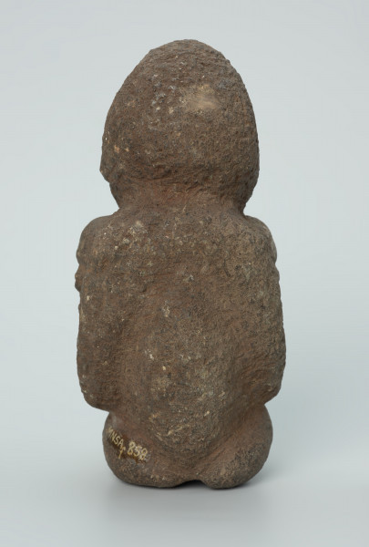 rzeźba; przedmiot obrzędowy; figura kultu zmarłych - Ujęcie z tyłu. Rzeźbiona w ziemistym steatycie siedząca postać kobiety z wyłaniającymi się symetrycznie zza jej boków dwoma ludzkimi głowami.