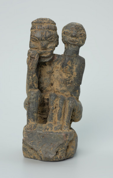 figura kultu przodków - Ujęcie z przodu. Rzeźbione w grafitowym kamieniu dwie złączone ze sobą postaci ludzkie w pozycji siedzącej. Stykają się ramionami, tułowiem oraz nogami. Jedna z nich – najprawdopodobniej mężczyzna - trzyma w prawej dłoni fajkę. Rzeźba pokryta jest piaskiem. Widoczne rysy, mikropęknięcia, malutkie otworki, przebarwienia oraz ślady klejenia w okolicach szyi.