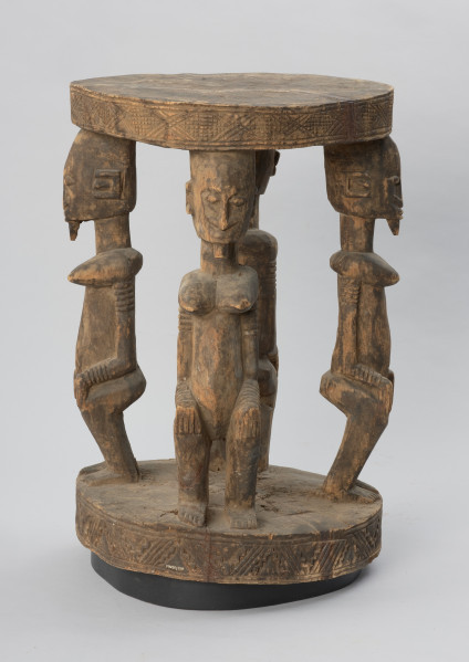 rzeźba - Ujęcie z tyłu z prawej strony. Drewniany stół. Okrągły blat połączony z podstawą o tym samym kształcie za pomocą czterech rzeźbionych postaci.