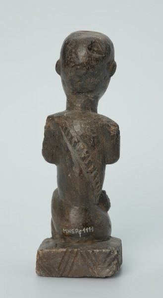 figura kultu przodków - Ujęcie z tyłu. Rzeźbiona w grafitowym kamieniu postać ludzka w pozycji siedzącej - najprawdopodobniej mężczyzna. Rzeźba ma charakterystyczne nacięcia - skaryfikacje wykonane na ramionach oraz plecach. Widoczne rysy i mikropęknięcia.