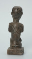 figura kultu przodków - Ujęcie z tyłu. Rzeźbiona w grafitowym kamieniu postać ludzka w pozycji siedzącej - najprawdopodobniej mężczyzna. Rzeźba ma charakterystyczne nacięcia - skaryfikacje wykonane na ramionach oraz plecach. Widoczne rysy i mikropęknięcia.