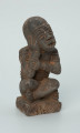 Ujęcie z przodu z prawej. Rzeźbiona w grafitowym kamieniu postać ludzka - najprawdopodobniej mężczyzna - w pozycji siedzącej z nogami skrzyżowanymi. Rzeźba ma charakterystyczne bransolety – dwie na ramionach oraz jedną na nadgarstku - które ozdobiono wzorem w postaci ukośnych linii. Widoczne ślady czerwonego piasku, rysy, mikropęknięcia oraz ubytek z tyłu głowy.