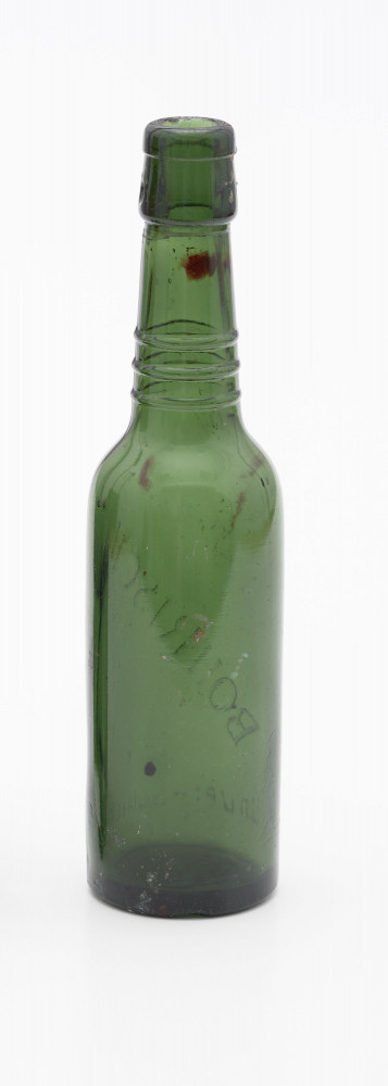szklana butelka z trzema wypukłymi pierścieniami na szyjce - Ujęcie z tyłu; Szklana butelka piwówka z grubego szkła butelkowego w ciemnym kolorze oliwkowo-zielonym. Szyjka wyodrębniona trzema wypukłymi pierścieniami liniowymi umieszczonymi w dolnej części szyjki. W górnej partii szyjki, poniżej wylewu zgrubienie z otworami na zamknięcie pałąkowe (brak). Ślady szwów bocznych pionowych. Dno nieco zagłębione.