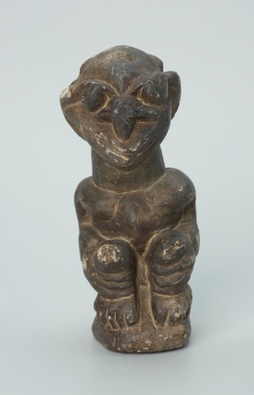 rzeźba; przedmiot obrzędowy; Figura kultu sił wegetacji - Ujęcie z przodu. Rzeźbiona w grafitowym steatycie siedząca postać ludzka o dwóch twarzach, z przodu i z tyłu głowy.