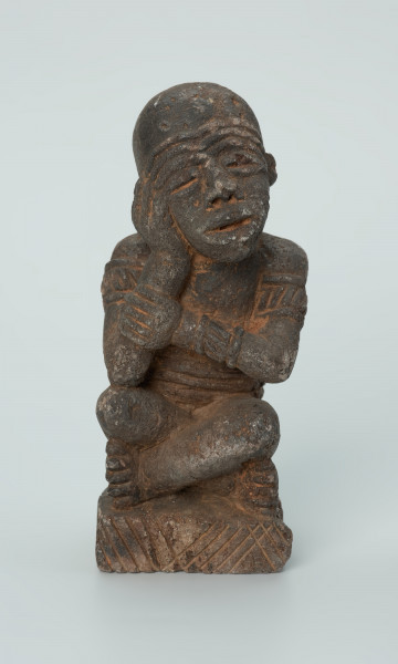 Ujęcie z przodu. Rzeźbiona w grafitowym kamieniu postać ludzka - najprawdopodobniej mężczyzna - w pozycji siedzącej z nogami skrzyżowanymi. Rzeźba ma charakterystyczne bransolety – dwie na ramionach oraz jedną na nadgarstku - które ozdobiono wzorem w postaci ukośnych linii. Widoczne ślady czerwonego piasku, rysy, mikropęknięcia oraz ubytek z tyłu głowy.