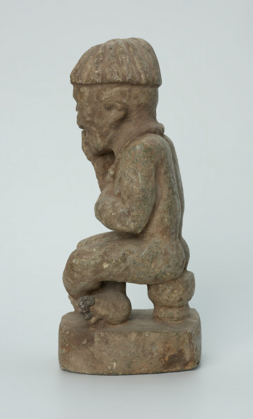 Ujęcie z boku z lewej. Rzeźbiona w biało-szarym kamieniu postać mężczyzny w pozycji siedzącej. Mężczyzna siedzi na okrągłym, malutkim stołku, ma charakterystyczne nakrycie głowy - czapkę modelowaną promieniście oraz naszyjnik. Widoczne rysy, mikropęknięcia i malutkie otworki.