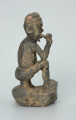 figura kultu przodków - Ujęcie z boku z prawej. Rzeźbione w grafitowym kamieniu dwie złączone ze sobą postaci ludzkie w pozycji siedzącej. Stykają się ramionami, tułowiem oraz nogami. Jedna z nich – najprawdopodobniej mężczyzna - trzyma w prawej dłoni fajkę. Rzeźba pokryta jest piaskiem. Widoczne rysy, mikropęknięcia, malutkie otworki, przebarwienia oraz ślady klejenia w okolicach szyi.