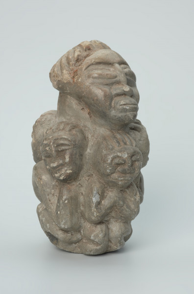 rzeźba; przedmiot obrzędowy; figura kultu zmarłych - Ujęcie z przodu z prawej strony. Rzeźba o gładkiej powierzchni w szarym steatycie, przedstawiająca postać ludzką o cechach kobiecych, otoczoną piątką mniejszych, dziecięcych sylwetek w różnych pozach.