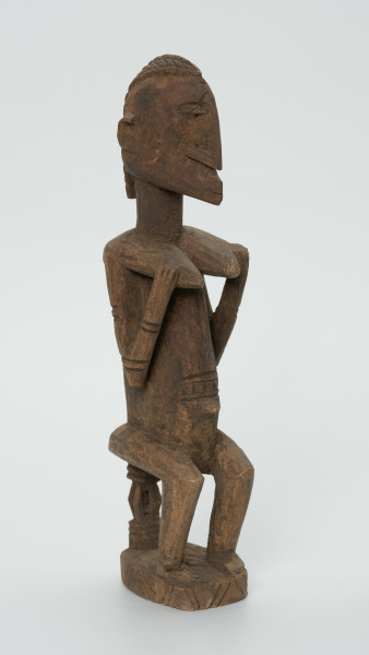 drewniana, rzeźbiona figura - Ujęcie z przodu, z lewej strony. Drewniana, rzeźbiona figura kobiety siedzącej na stołku.