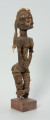 drewniana figura - Ujęcie z przodu, z prawej strony. Drewniana, rzeźbiona postać kobiety, która wokół nóg i rąk ma obwiązane wąskie paski skóry. Do głowy przymocowany jest pasek bawełny z naszytymi muszelkami kauri.