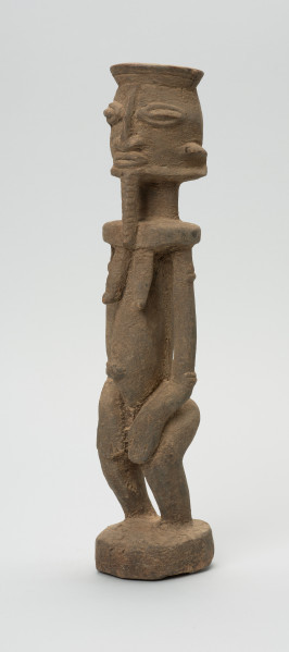 rzeźbiona figura - Ujęcie z przodu, z lewej strony. Drewniana, rzeźbiona postać, z zaznaczonymi jednocześnie cechami płciowymi męskimi i żeńskimi. Wyodrębnione cechy męskie to broda, żeńskie: piersi.